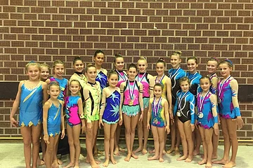 Gruppenfoto von 20 jungen Turnerinnen in bunten Turnanzügen teilweise mit Medaillen um den Hals vor einer braunen Wand stehend.