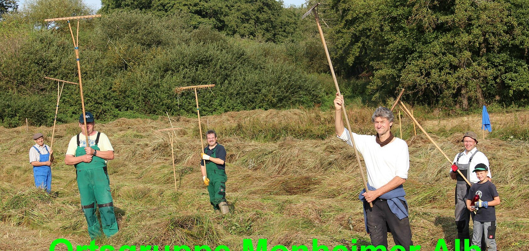 Sechs Personen in Arbeitshosen stehen bei Tageslicht auf einer Wiese vor Bäumen mit hochgehobenen Rechen in den Händen. Auf dem Foto der Text „Bund Naturschutz in Bayern e.V. - Ortsgruppe Monheimer Alb“ in grün.