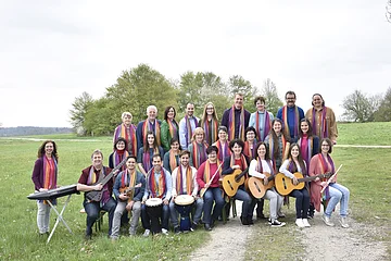 Gruppenfoto von 28 Personen mit Musikinstrumenten und bunten Schals um den Hals auf einer Wiese vor Bäumen bei bewölktem Himmel.