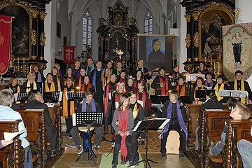 Chor mit bunten Schals singt und musiziert in einer Kirche vor Publikum auf braunen Holzbankreihen.