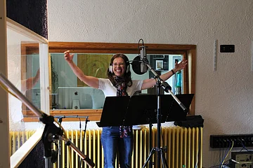 Frau mit Kopfhörern steht vor einem Mikrofon und Notenständer in Tonstudio mit ausgestreckten Armen.