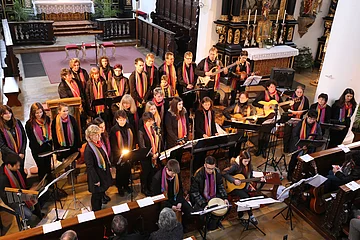 Chor mit Instrumenten und bunten Schals singt und spielt in einer Kirche vor Publikum.