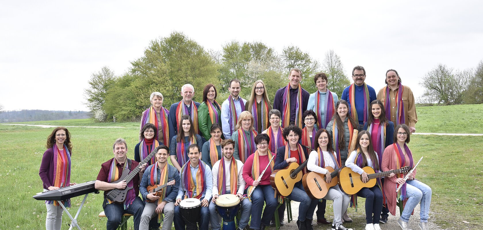 Gruppenfoto von 28 Personen mit Musikinstrumenten und bunten Schals um den Hals auf einer Wiese vor Bäumen bei bewölktem Himmel.
