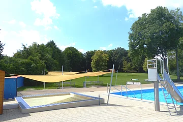 Außenbereich eines Freibads mit Planschbecken für Kinder, einem Schwimmerbecken, Rutsche aus Metall und gespannten Sonnensegeln bei Sonnenschein. Im Hintergrund grüne Wiese und Bäume.
