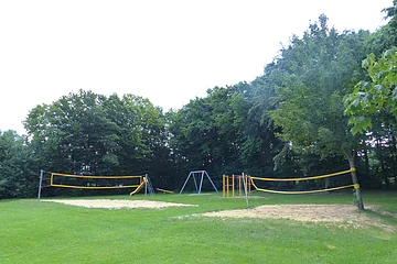 Spielplatz mit zwei gelben Volleyballnetzen, einer Rutsche, einer Schaukel und Klettergerüst vor Baumreihe auf Grasfläche, bei Tageslicht.