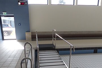 Schwimmbeckenbereich in einem Hallenbad mit leerem Schwimmbecken mit Rampe aus Metall, die in das Becken führt und einer gefließten Treppe. Drei Fenster oben an der Wand und eine Ausgangstür links davon im Hintergrund.