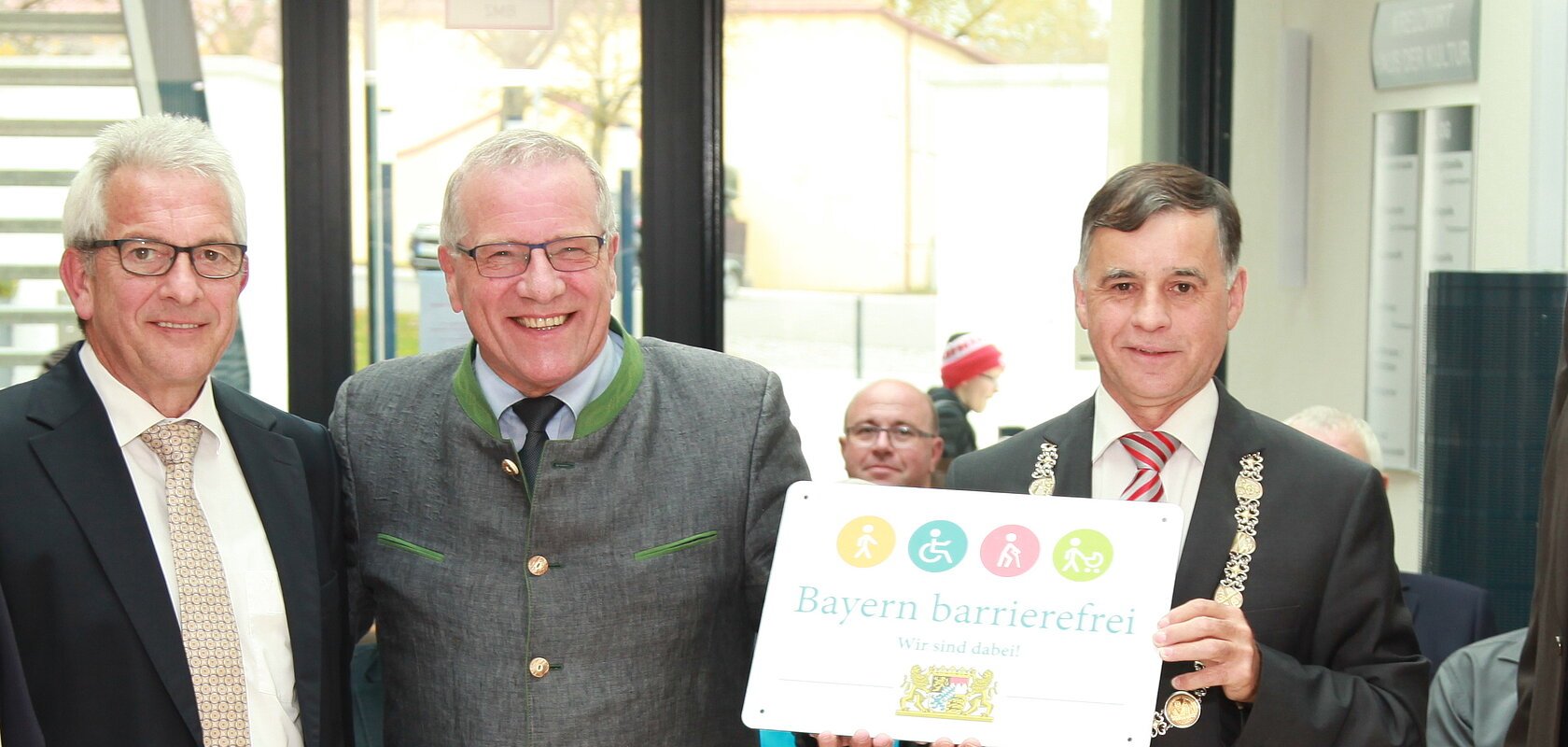 Fünf Männer in Anzügen stehen vor Stühlen in einem hellen Raum nebeneinander, einer hält ein weißes Schild mit der Aufschrift „Bayern barrierefrei“, darauf oberhalb der Schrift verschiedene bunte Symbole und darunter das Wappen von Bayern. Im Hintergrund sitzen weitere Menschen auf Stühlen vor einem Glasausgang.