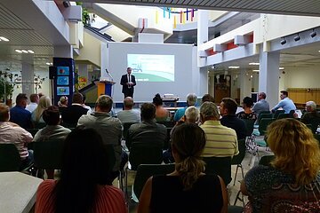 Publikum sitzt auf Stühlen in einem Raum und hört einem Mann im Anzug neben einem Rednerpult zu, der vor einer Leinwand eine Präsentation hält. Oberhalb weitere Stockwerke des Gebäudes mit Treppen aus Beton zu sehen, sowie bunte Dekorationen.