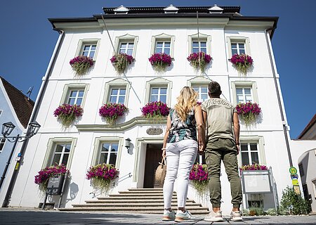 Paar, Mann und Frau von hinten vor einem weißem Gebäude mit Blumen in Blumenkästen an den Fenstern, schauen auf das Gebäude bei klarem Himmel. Eine Infovitrine auf der rechten Seite am Gebäude.
