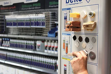 Hand hält Musterstück an ein Display mit Klebstoffproben und ausgestellten Materialien in Regalen eines Fachgeschäfts.