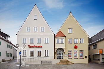 Zwei nebeneinanderstehende mehrstöckige Gebäude mit spitzen Dächern, auf dem Linken Gebäude in beige der Schriftzug "Sparkasse" in roter Farbe, auf dem rechten Gebäude das Sparkassen-Logo ein Großes S in rot mit einem roten Punkt darüber. Vor den zwei Gebäuden eine Treppe und eine barrierefreie Auffahrt, auf einem gepflastertem Platz bei blauem Himmel. Links und rechts weitere Gebäude.