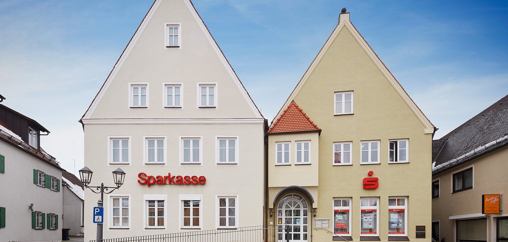 Zwei nebeneinanderstehende mehrstöckige Gebäude mit spitzen Dächern, auf dem Linken Gebäude in beige der Schriftzug "Sparkasse" in roter Farbe, auf dem rechten Gebäude das Sparkassen-Logo ein Großes S in rot mit einem roten Punkt darüber. Vor den zwei Gebäuden eine Treppe und eine barrierefreie Auffahrt, auf einem gepflastertem Platz bei blauem Himmel. Links und rechts weitere Gebäude.