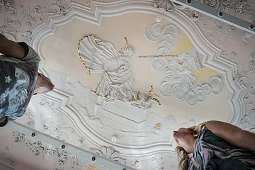 Stuckdecke im Monheimer Rathaus mit barocker farbiger Stuckreliefdarstellung und zwei Personen, die von unten darauf schauen.