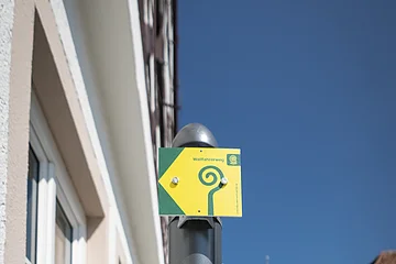 Drei leuchtend gelbe Schilder am grauen Pfosten in einer Altstadt bei blauem Himmel. Zwei Schilder zu Wanderwegen mit der Nummern 5 und 6 und ein Pilgerweg-Symbol für den Wallfahrerweg, mit dem dazugehörigen spiralförmigen Symbol.