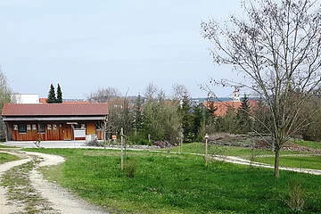Zwei Schotterwege links und rechts führen durch grüne Wiese zu einem Holzhaus mit roten Dach, Bäume und Dorf mit Kirchturm bei bewölktem Himmel im Hintergrund.