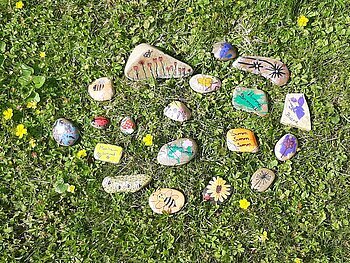 Mehrere bemalte Steine mit Blumen, Insekten und Texten liegen auf grünem Gras mit gelben Blumen.