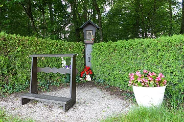 Begrenzter Gebetsplatz mit Holzkneeler auf Schottersteinen, Blumen und kleinem Bildstock vor grüner Hecke im Wald.