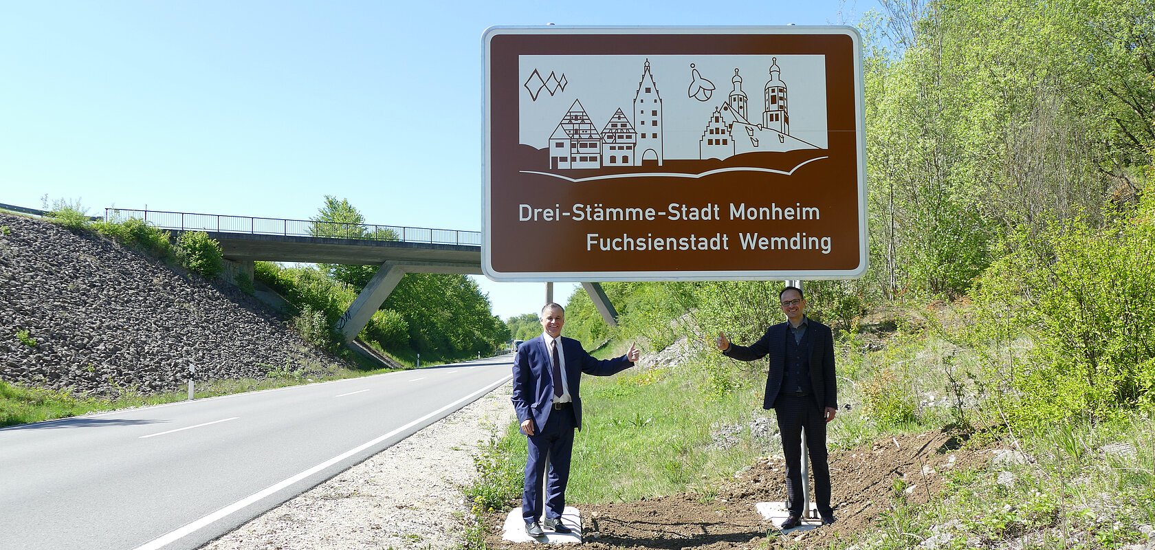 Zwei Männer in Anzügen stehen an einer Verkehrsstraße vor einem braunen Schild mit der Aufschrift „Drei-Stämme-Stadt Monheim Fuchsienstadt Wemding“. Über dem Text eine Abbildung mit den Umrissen zweier verschiedenen Städte.