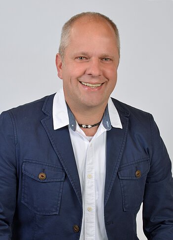 Porträt eines lächelnden Mannes mit kurzem Haar, weißem Hemd, dunkelblauem Sakko und einer Halskette, vor hellem Hintergrund.