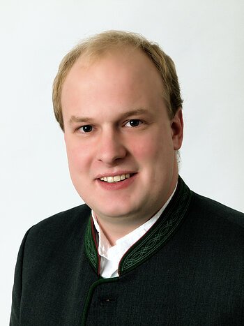 Porträt eines lächelnden Mannes mit kurzem blondem Haar in traditioneller dunkelgrüner Trachtenjacke vor weißem Hintergrund.