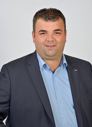 Porträt eines Mannes mit kurzen dunklen Haaren, blauem Hemd und dunklem Sakko, vor hellgrauem Hintergrund.