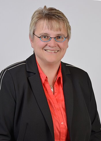 Porträt einer lächelnden Person mit kurzen blonden Haaren, Brille, rotem Hemd und schwarzem Blazer, vor hellem Hintergrund.