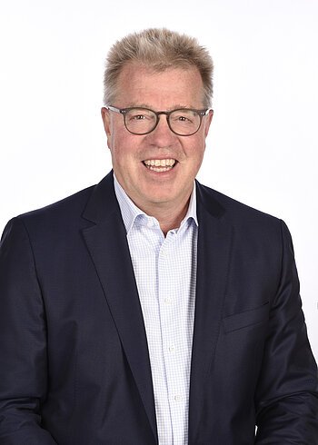 Porträt eines lächelnden Mannes mit Brille, hellblondem Haar, weißem gestreiften Hemd und dunklem Sakko, vor weißem Hintergrund.