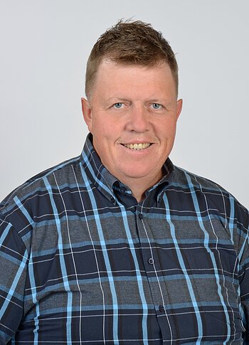 Porträt eines lächelnden Mannes mit kurzem Haar und kariertem Hemd, vor hellem Hintergrund.