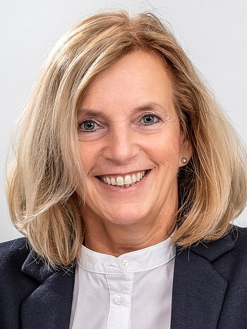 Porträt einer lächelnden Frau mit schulterlangem blondem Haar, weißer Bluse und dunklem Blazer vor hellem Hintergrund.