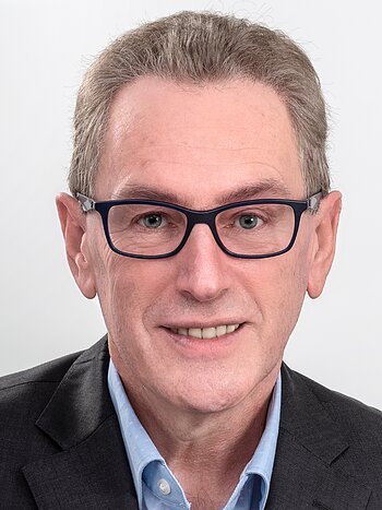 Porträt eines älteren Mannes mit dunkelblondem Haar, Brille, blauem Hemd und dunklem Sakko vor hellem Hintergrund.