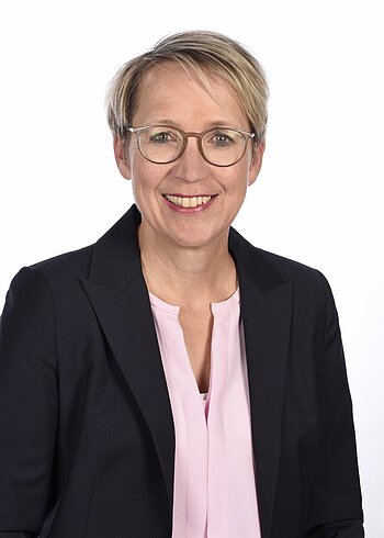 Porträt einer Frau mit kurzen blonden Haaren, Brille, rosa Bluse und dunklem Blazer, vor weißem Hintergrund.