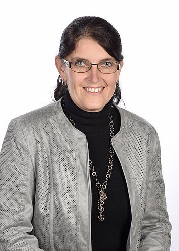 Porträt einer lächelnden Frau mit Brille, dunklen Haaren, grauer Jacke, schwarzem Rollkragenpullover und Kette, vor weißem Hintergrund.