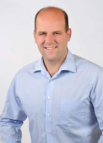 Porträt eines lächelnden Mannes mit hellblauem Hemd vor weißem Hintergrund.