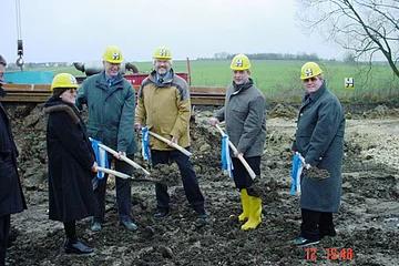 Fünf Personen mit gelben Helmen und Schaufeln stehen auf einer Baustelle mit Erde und Wiese im Hintergrund bei bewölktem Himmel.