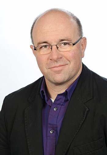 Porträt eines Mannes mit Brille, schwarzem Sakko und lila Hemd vor weißem Hintergrund.