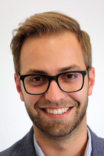 Portrait eines lächelnden Mannes mit Brille, in ein Sakko und ein Hemd gekleidet, vor einem weißem Hintergrund.