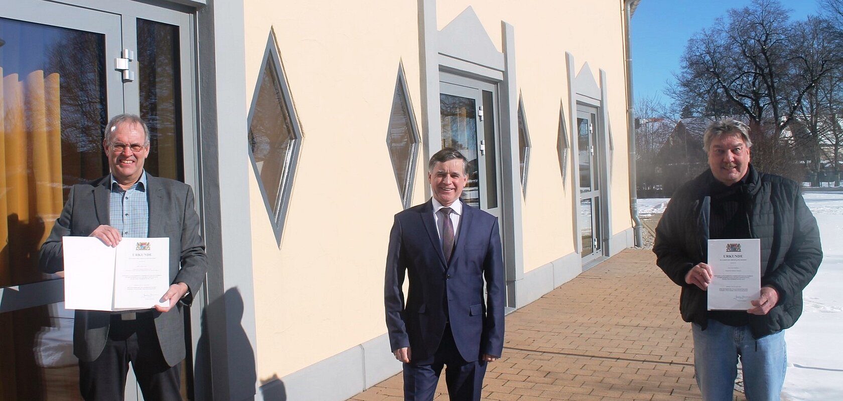 Drei Männer stehen vor einem hellen Gebäude auf einem gepflastertem Platz, zwei halten Urkunden in den Händen, Schnee rechts im Bild liegt auf dem Boden.