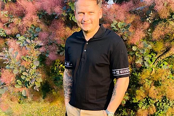 Mann mit schwarzem Poloshirt und beigen Shorts steht auf Rasen vor buschigem Baum mit rosa Blüten.