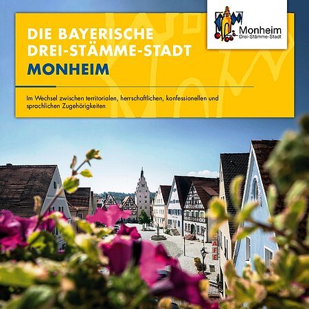 Titelblatt eines Folders zur Stadt Monheim mit dem Titel "Die bayerische Drei Stämme-Stadt Monheim" in weißer Schrift auf gelben Hintergrund. Auf dem Foto der Blick auf den historischen Marktplatz mit Fachwerkhäusern und einem Turm in Monheim, davor unscharf pinke Blumen, bei blauem Himmel.