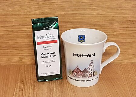 Geschenkidee der Stadt Monheim: eine weiße Tasse mit Monheim-Motiv und der Aufschrift "Monheim - Die Drei-Stämme-Stadt" rechts im Bild. Links davon eine Teeverpackung mit der Mischung und dem Text „Monheimer Früchtekorb“ stehen vor einer Holzoberfläche.