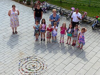 Gruppe von 8 Kindern und 4 Erwachsenen steht auf gepflastertem Platz neben einer Spirale aus bemalten Steinen. Hinter ihnen langer Fahrrad-Abstellplatz mit Laufrädern und Fahrrädern.