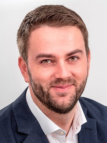 Porträt eines lächelnden Mannes mit kurzem braunem Haar, Bart, blauem Sakko und weißem Hemd, vor hellem Hintergrund.