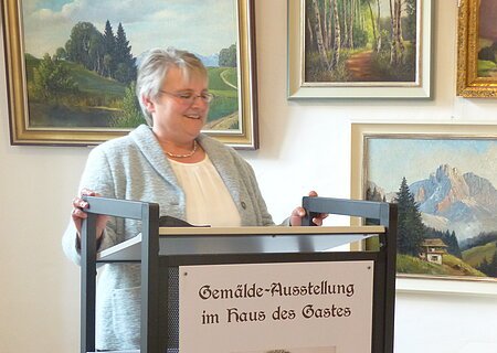 Frau steht an einem Rednerpult mit einem am Pult angebrachtem Plakat zur Gemälde-Ausstellung von Hans Mayer im Haus des Gastes. Hinter Ihr an der weißen Wand sind Gemälden von Landschaften zu sehen.