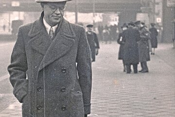 Mann mit Hut und Mantel geht an Leine mit großem schwarzen Hund auf gepflastertem Platz, Personen im Hintergrund.