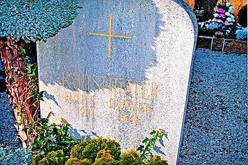 Grabstein mit der goldener Aufschrift „Kunstmaler Hans Mayer 1901 - 1958“ und einem goldenem Kreuz, umgeben von Pflanzen. Im Hintergrund Schottersteine und weiters Grab.