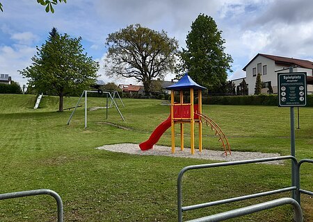 Spielplatz mit Rutsche, Klettergerüst, Seilbahn, Schaukel und Hinweisschild auf einer Wiese vor Wohnhäusern bei bewölktem Himmel.