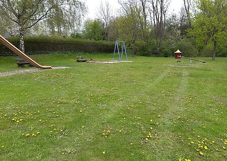 Spielplatz mit Rutsche, Schaukel, Wippe, zwei Bänken und Wiese mit gelben Blumen. Im Hintergrund sind Bäume bei bewölktem Himmel.