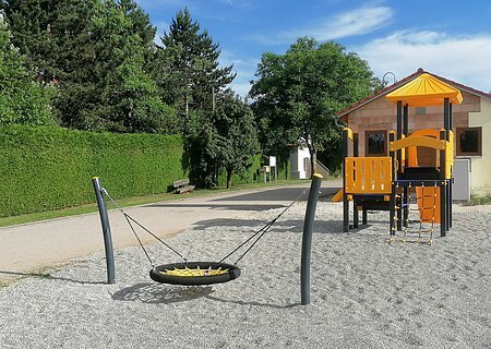 Spielplatz mit Schottersteinen einer schwarz-gelber Nestschaukel und schwarz-orangem Klettergerüst vor einem unverputzten Haus und Bäumen links davon, bei sonnigem Himmel.