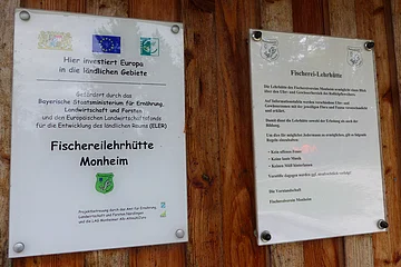 Zwei Informationsschilder befestigt an einer Holzwand zur Fischereilehrhütte Monheim mit Förderhinweisen und Nutzungsregeln der Fischereilehrhütte.