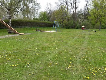 Spielplatz mit Rutsche, Schaukel, Wippe, zwei Bänken und Wiese mit gelben Blumen. Im Hintergrund sind Bäume bei bewölktem Himmel.